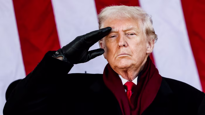 Donald Trump salutes, wearing a black glove and red scarf, in front of an American flag backdrop.