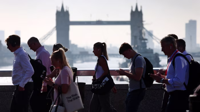 Commuters walk across London Bridge in the morning with Tower Bridge visible in the background. Some look at phones or wear headphones.