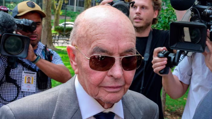 Joe Lewis, wearing sunglasses and a suit, is surrounded by members of the press with cameras outside.