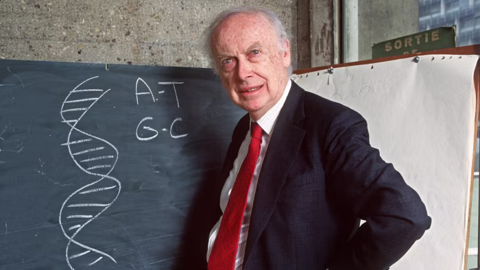 James Dewey Watson stands in front of a blackboard with a chalk drawing of a DNA double helix and base pair letters A-T, G-C.