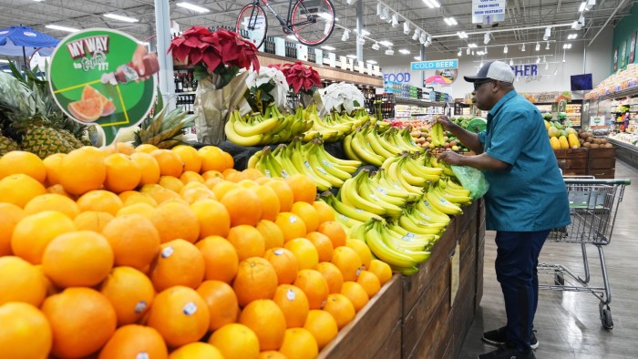A man selects bananas in the produce section of a supermarket, with oranges and pineapples displayed nearby.