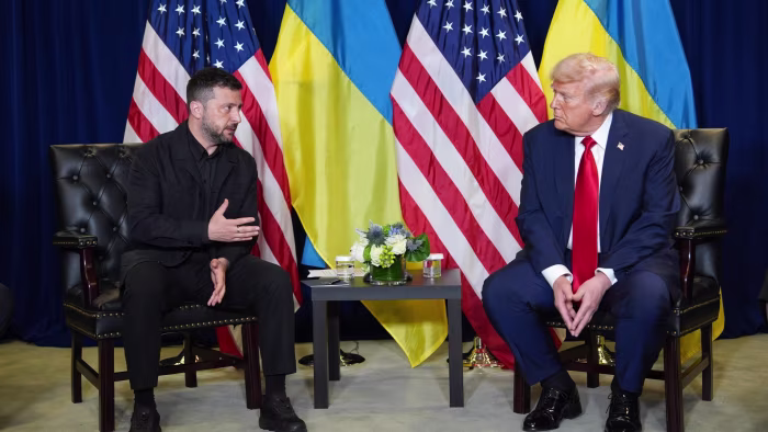 Volodymyr Zelenskyy gestures while speaking with Donald Trump as they sit across from each other, with U.S. and Ukrainian flags behind them.