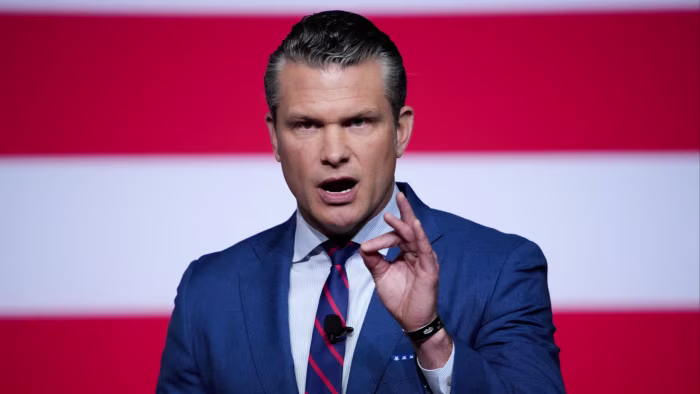 Pete Hegseth speaks with a serious expression, gesturing with his hand, in front of a backdrop with red and white stripes.