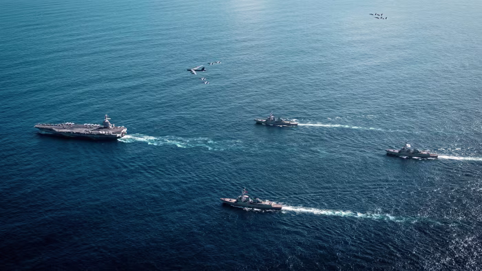 Four U.S. Navy warships, including the USS Gerald R. Ford, sail in formation below several F/A-18E/F Super Hornets and a B-52 bomber.