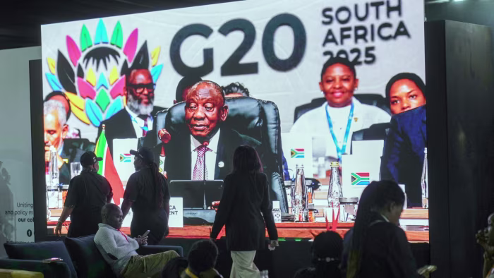 Cyril Ramaphosa is shown on a large screen holding a wooden gavel as he closes the G20 leaders' summit; people walk by in front of the screen.