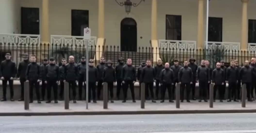 A group of black-clad men stand with their arms by their side in front of the NSW Parliament.