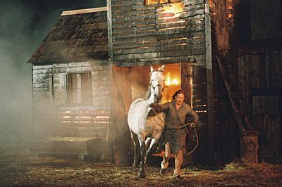 A man runs, guiding a white horse away from a burning wooden building engulfed in flames. It is nighttime.