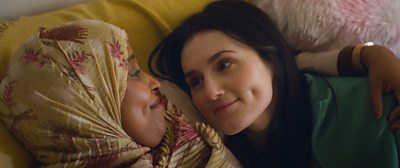 Two women with their heads resting on a cushion smiling at each other. One wears a pretty headscarf with pink floral detail.