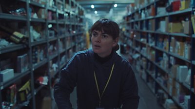 A woman in a hoodie, wearing a yellow lanyard walks down an aisle. Either side of the aisle are storage shelves full of box files