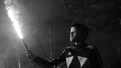 A blacka nd white image of a young boy in sports wear holding a lit flare in his right hand. There are trees behind him.