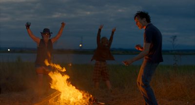 Three people dancing on grass, around a camp fire, next to a body of water