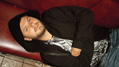 A young man in a black hoodie lies with his arms folded on some red leather seating, looking at the camera which is above him.