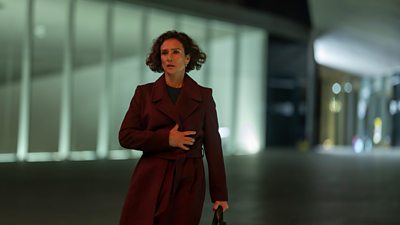 A woman walks at night holding her coat closed and looking concerned