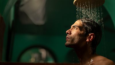 A man stands beneath a running shower head looking towards a light that shines on his face
