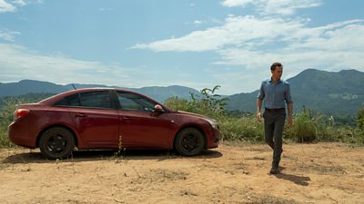 A man in jeans and a shirt walks past a red car on a hillside, set against a backdrop of mountains