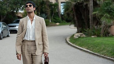 A man in a light linen suit, white shirt and sunglasses walks down a residential street past cars and palm trees, holding a satchel/briefcase