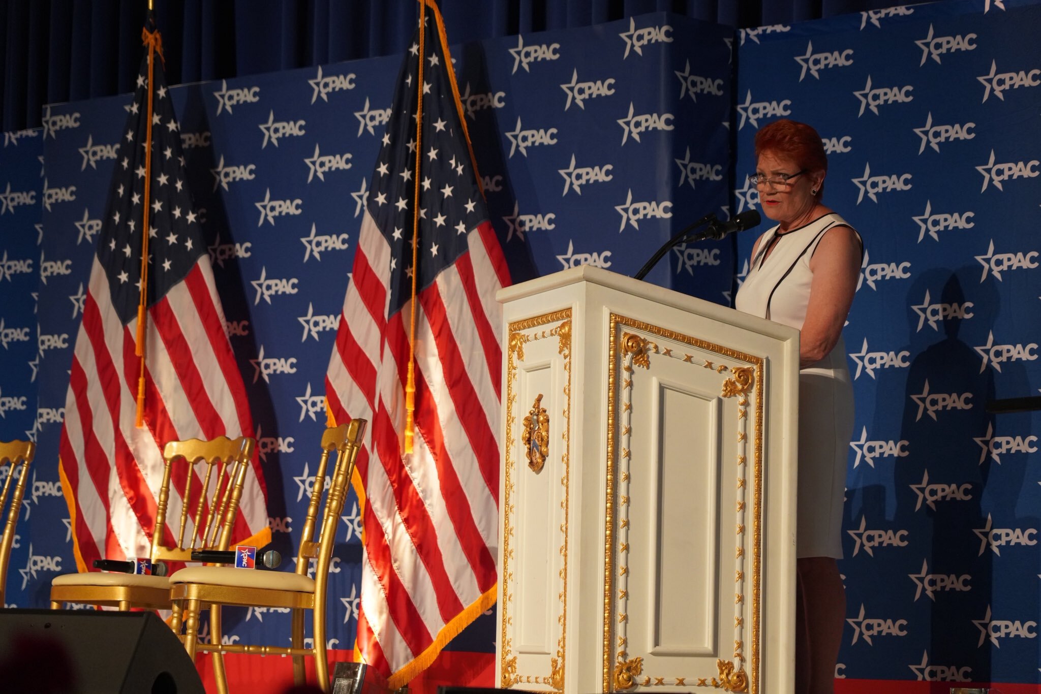 A woman, with short red hair, in a white dress, stands at a white lectern with gold trimmings on it. American flags are to the left of the image.
