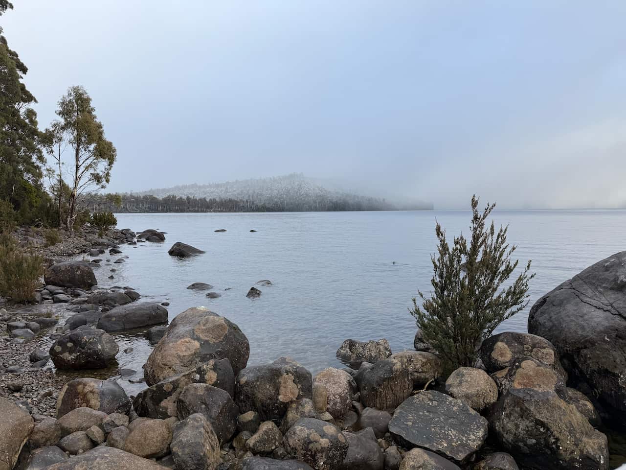 A rocky shoreline and lake. In the distance, clouds and snow can be seen on hills. 