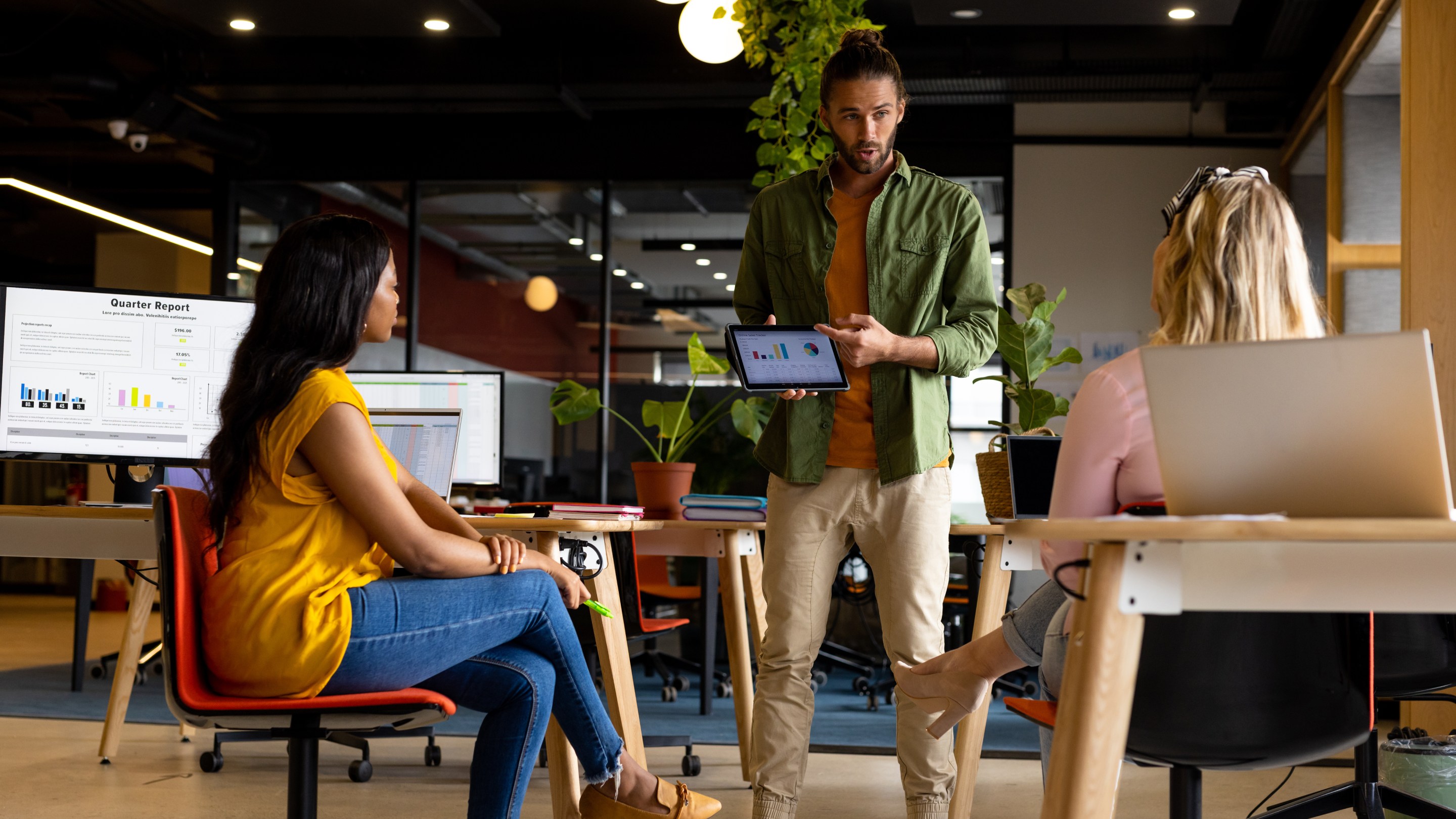 Three colleagues in a modern office during an informal presentation, with one person standing and showing charts on a tablet while two others sit at desks with laptops and a large screen displaying a report.