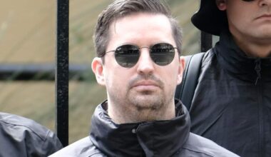 A young white man with brown hair, round sunglasses and a dark parka jacket. He's standing among a group of other men in similar clothing.