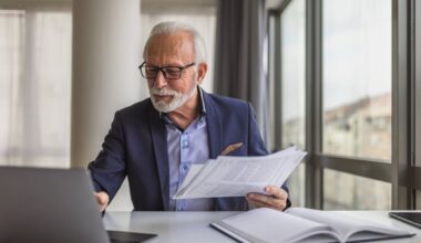Person in office, holding documents and looking at laptop.