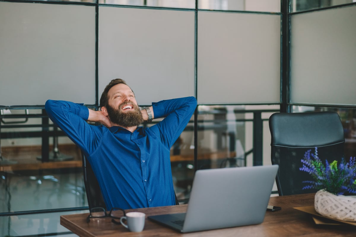 Professional leaning back in chair in office, smiling and relaxing at a desk with coffee and laptop.