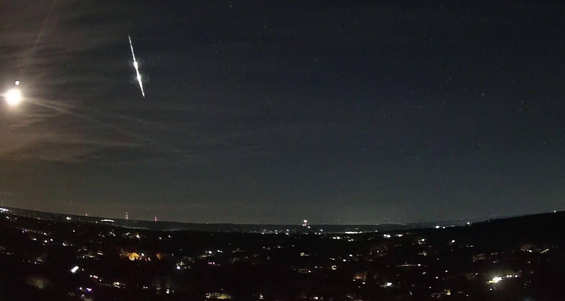 A bright fireball meteor is shown streaking earthward through the night sky over the lights of a town. The bright reflected light of the moon is visible to its left.