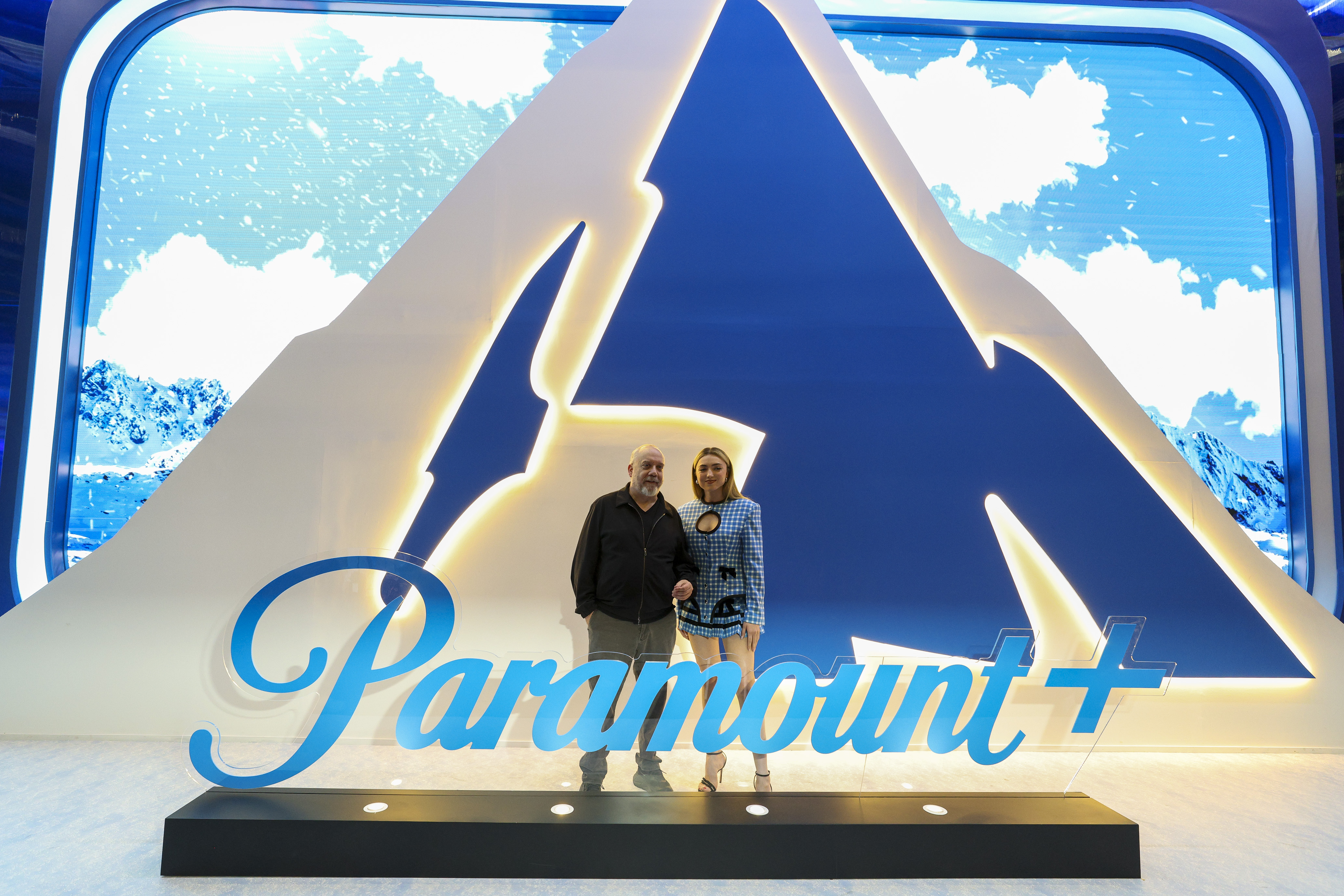 Paul Giamatti and Peyton List posing in front of a large, illuminated Paramount+ logo at CCXP Sao Paulo 2025.