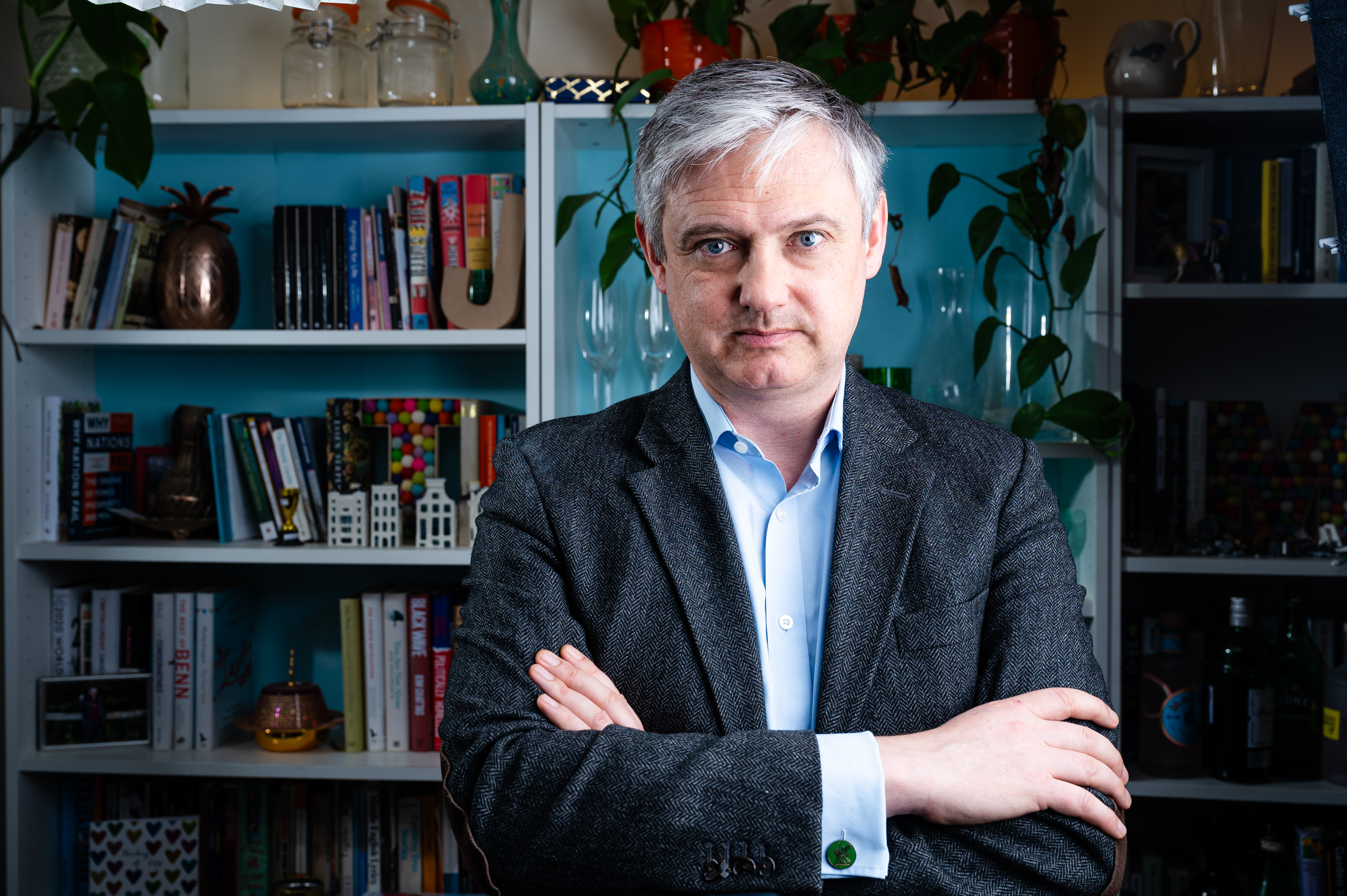 Lord Walney (John Woodcock) poses at home, wearing a tweed jacket and light blue shirt, against a bookshelf filled with books and decorative items.