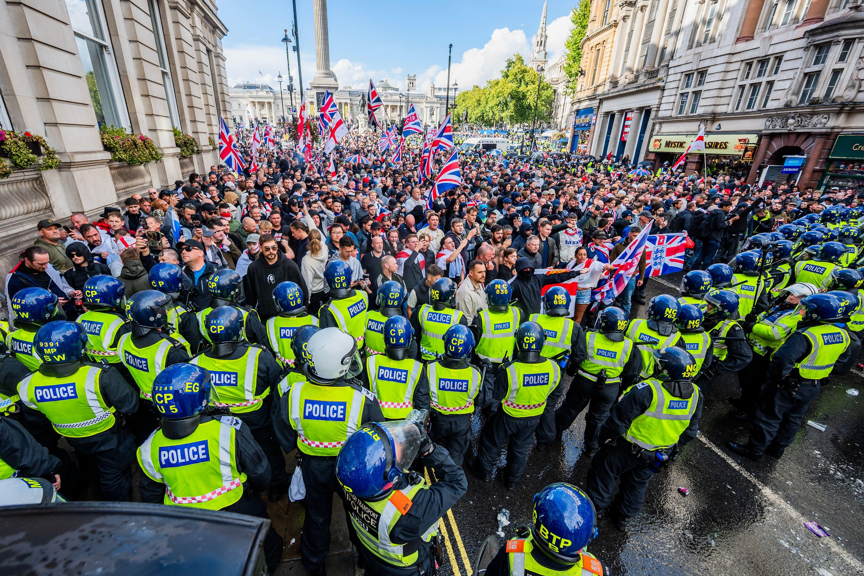 Police officers in riot gear facing a crowd of right-wing protesters in London, many holding Union Jack flags.