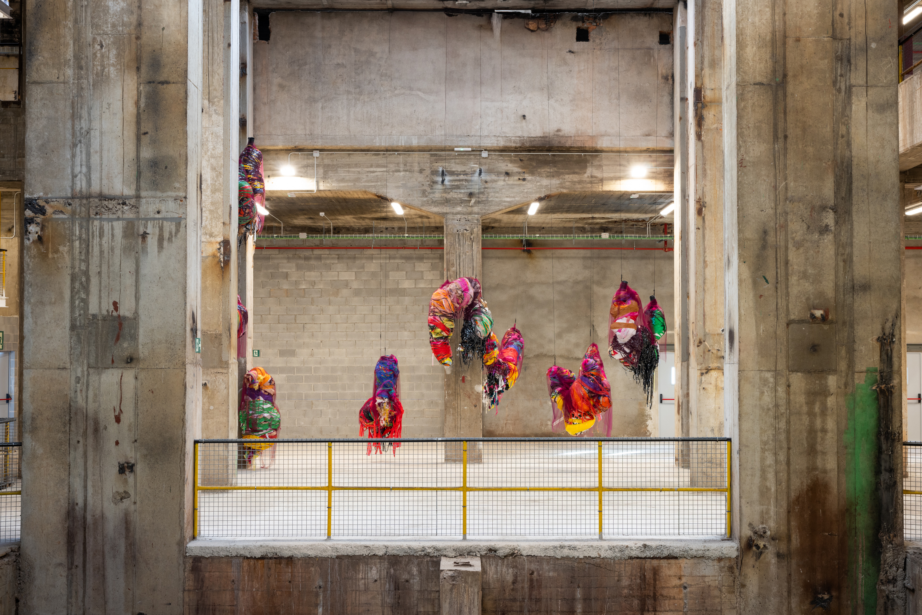 Hanging Sculpture 1 to 10 by Nnena Kalu, featuring colorful, abstract forms made from various materials, suspended in an industrial concrete space.