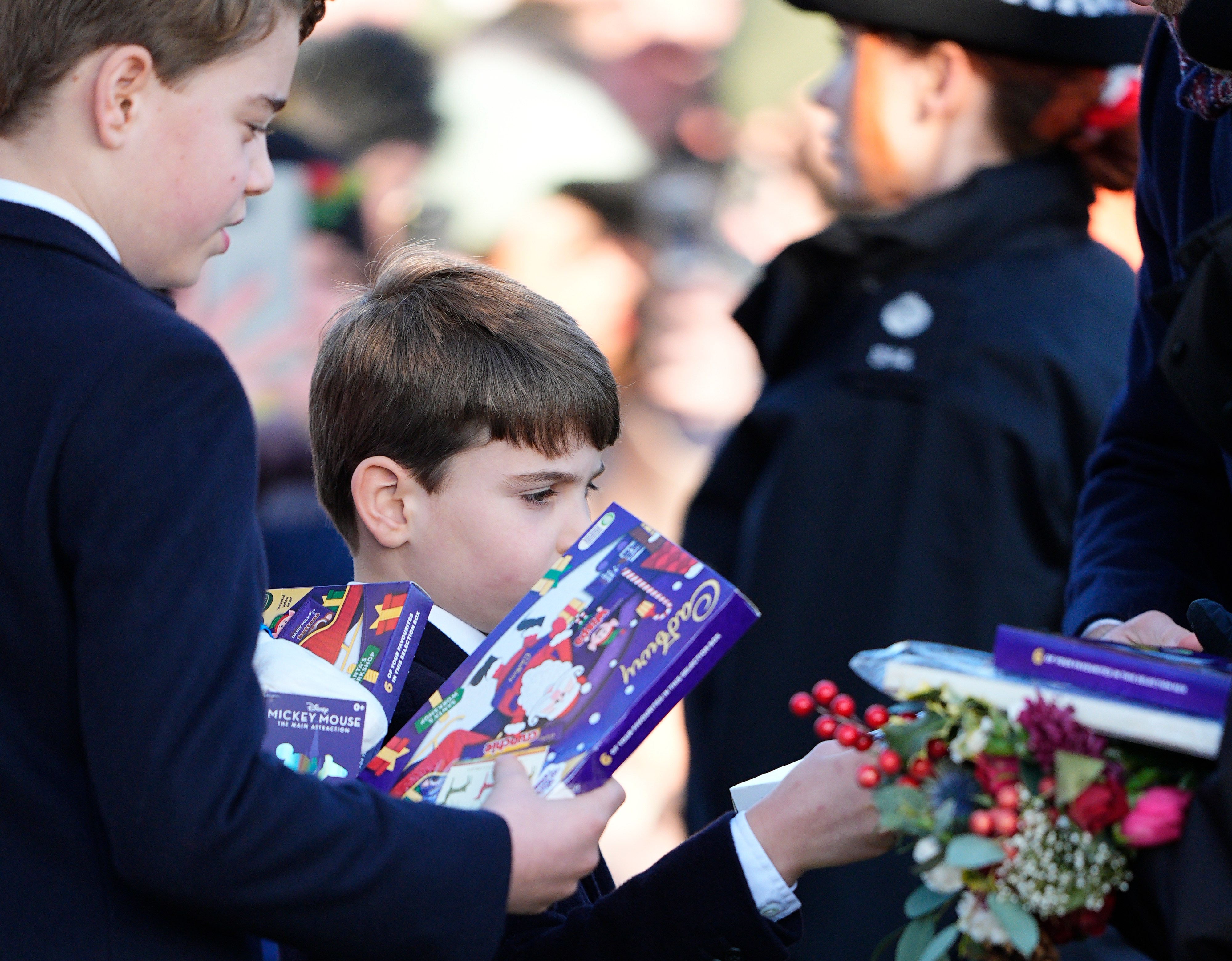 Prince Louis holds a Cadbury selection box, a Mickey Mouse toy, and a small bouquet of flowers.