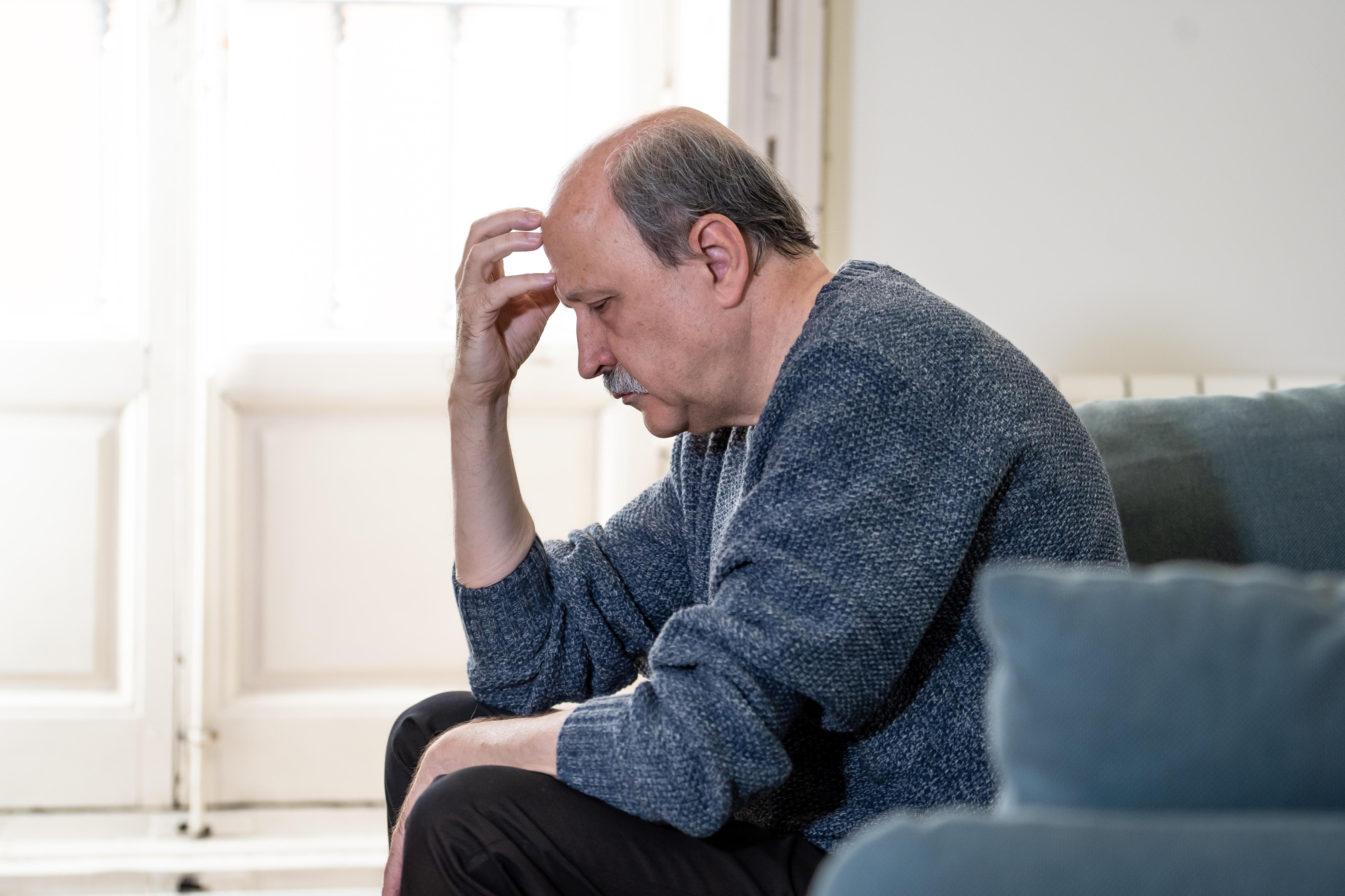 A sad and desperate elderly man with his head in his hand, sitting on a couch.