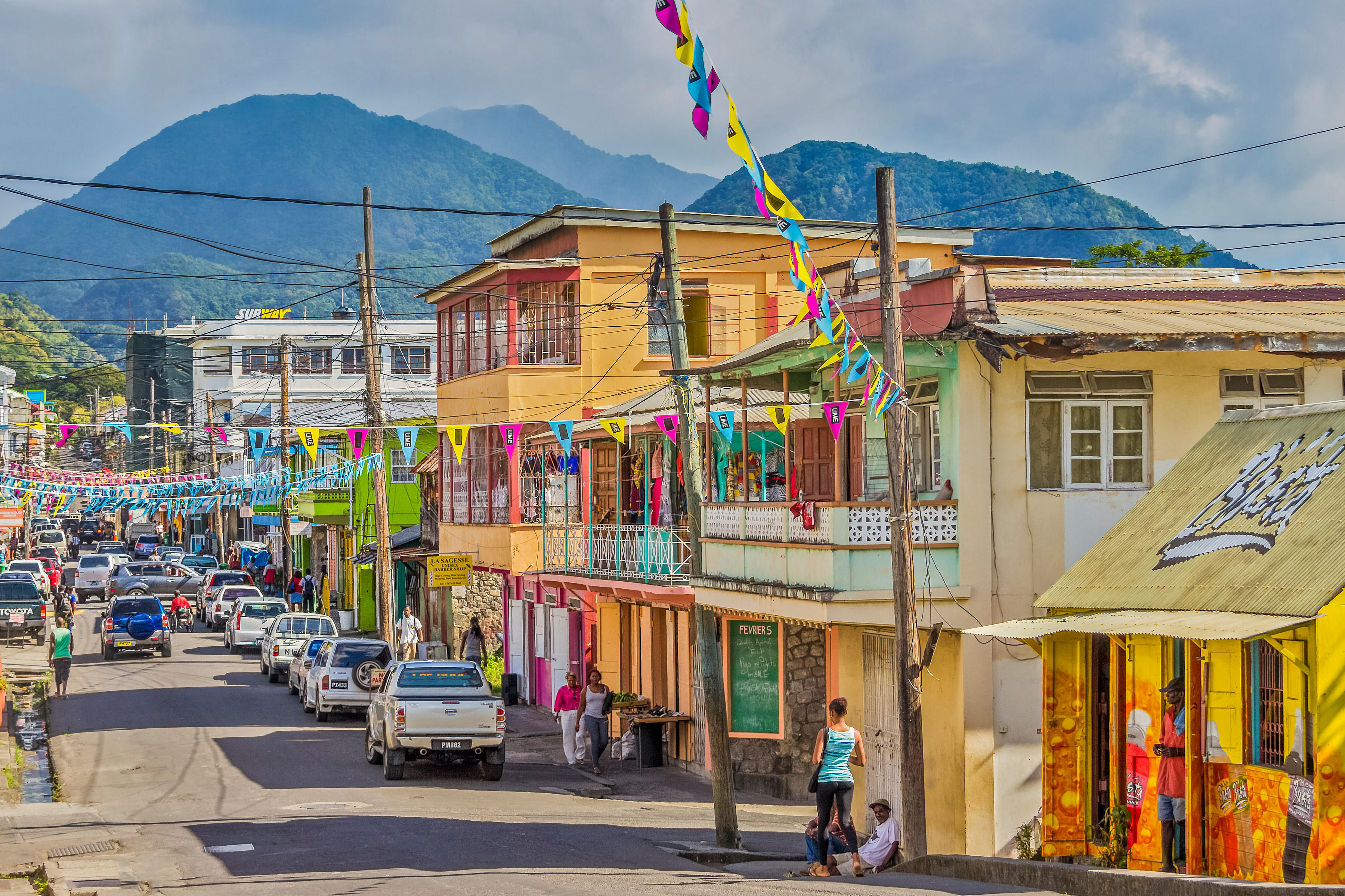 A busy road in Roseau, Dominica, West Indies, with colorful buildings, vehicles, and pedestrians, and mountains in the background.
