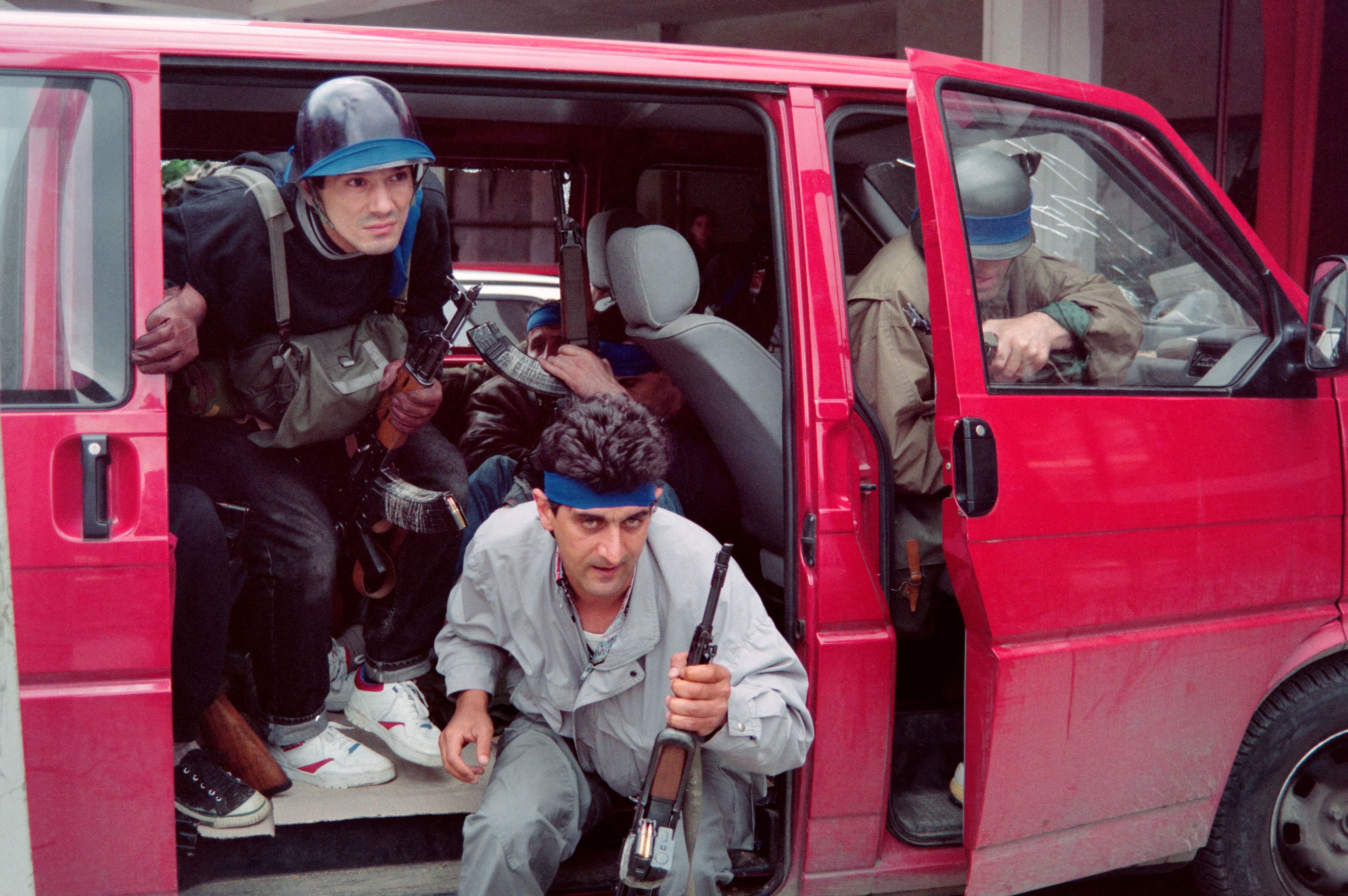 Bosnian fighters exiting a red civilian van, carrying rifles.