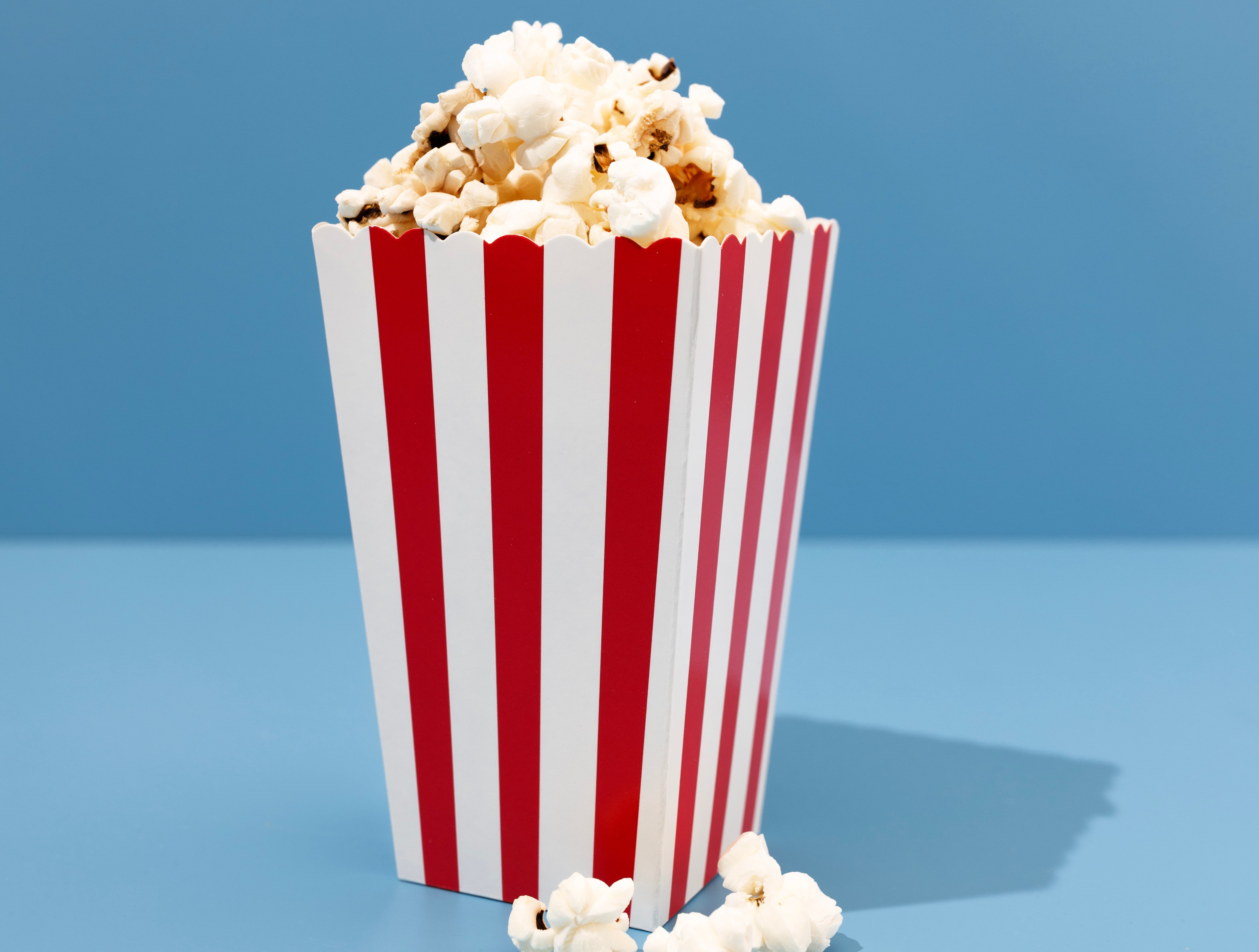 A red and white striped popcorn bucket overflowing with popcorn, set against a blue background.