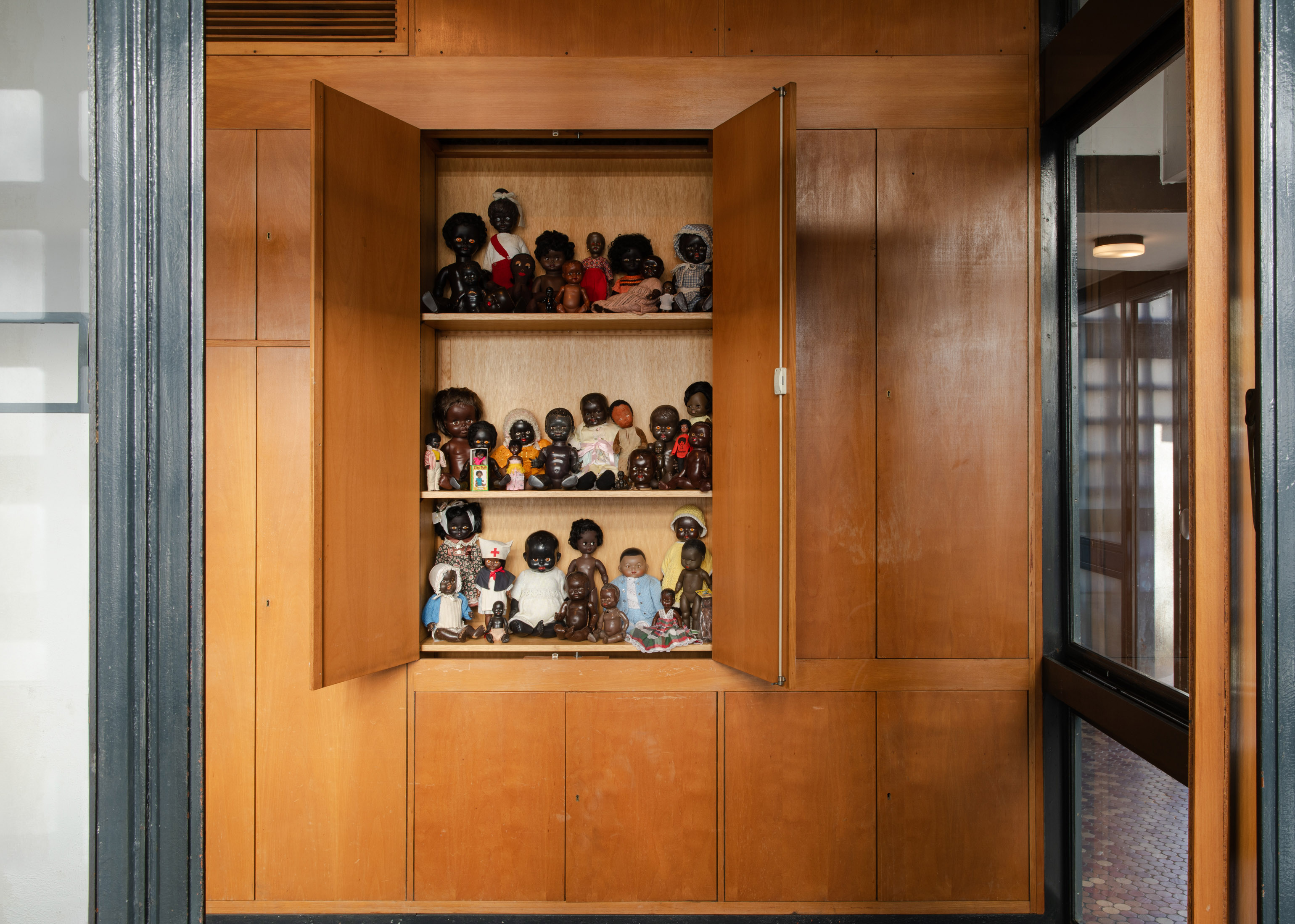 An open wooden cabinet filled with rows of diverse Black dolls, part of the artwork "AS OPPOSED TO THE TRUTH" by Rene Matic.