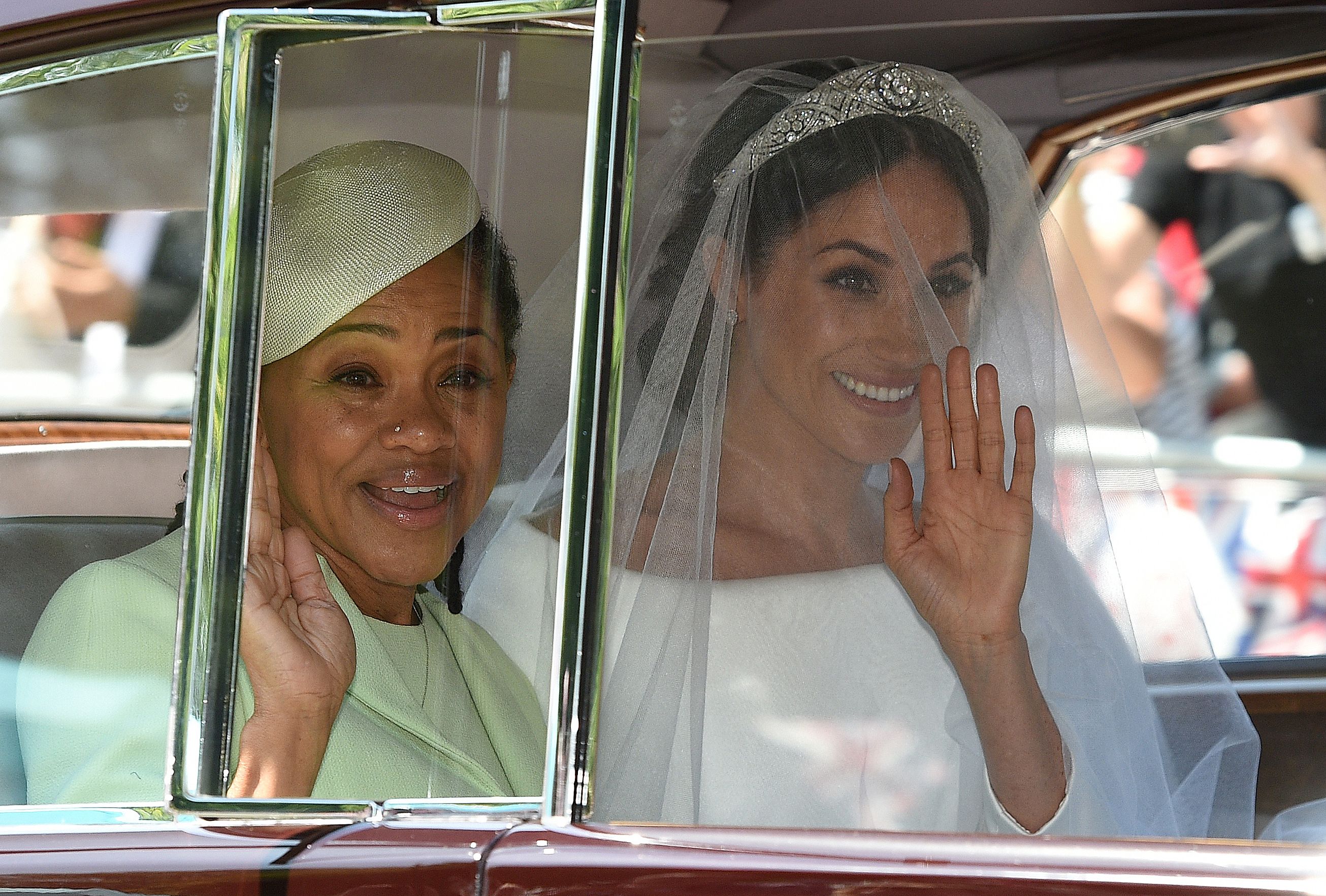 Meghan Markle in a wedding dress and veil and her mother, Doria Ragland, in a light green hat and dress, both waving from inside a car.