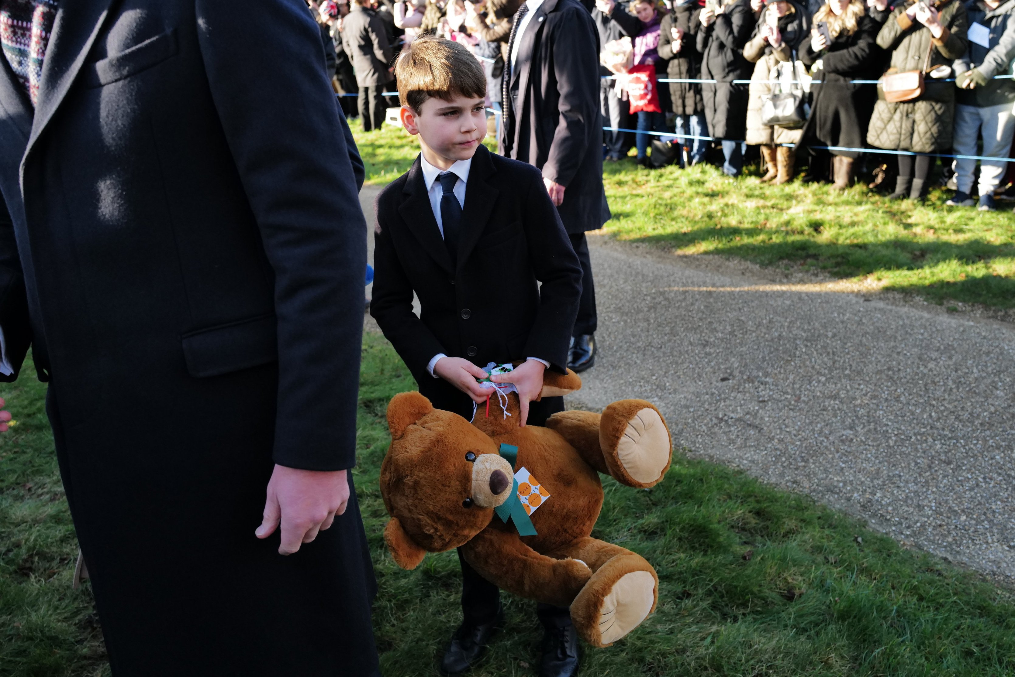 A young boy in a suit holds a teddy bear as members of the public watch the Royal family attend a Christmas service.