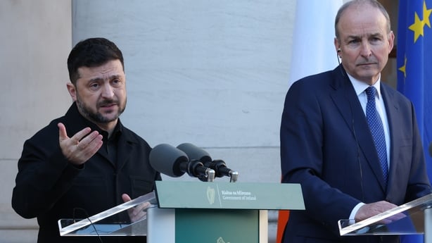 A man with short dark hair stands at a podium with two microphones. He is wearing dark clothes. A taller balding man stands at a podium beside him. He is wearing a navy suit and tie with a white shirt. The background is white.
