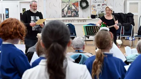 Jo Hislop The backs of the heads of the primary school children who are wearing blue and white school uniforms. In front of them, Sara Dennis and Bob Beagrie and performing. Ms Dennis has short, blonde hair and is playing a small guitar. Mr Beagrie is wearing a black jumper and has grey short hair and a grey beard. They are standing in a room with patterned, beige wallpaper. There are two posters on the wall and white double doors to the left.
