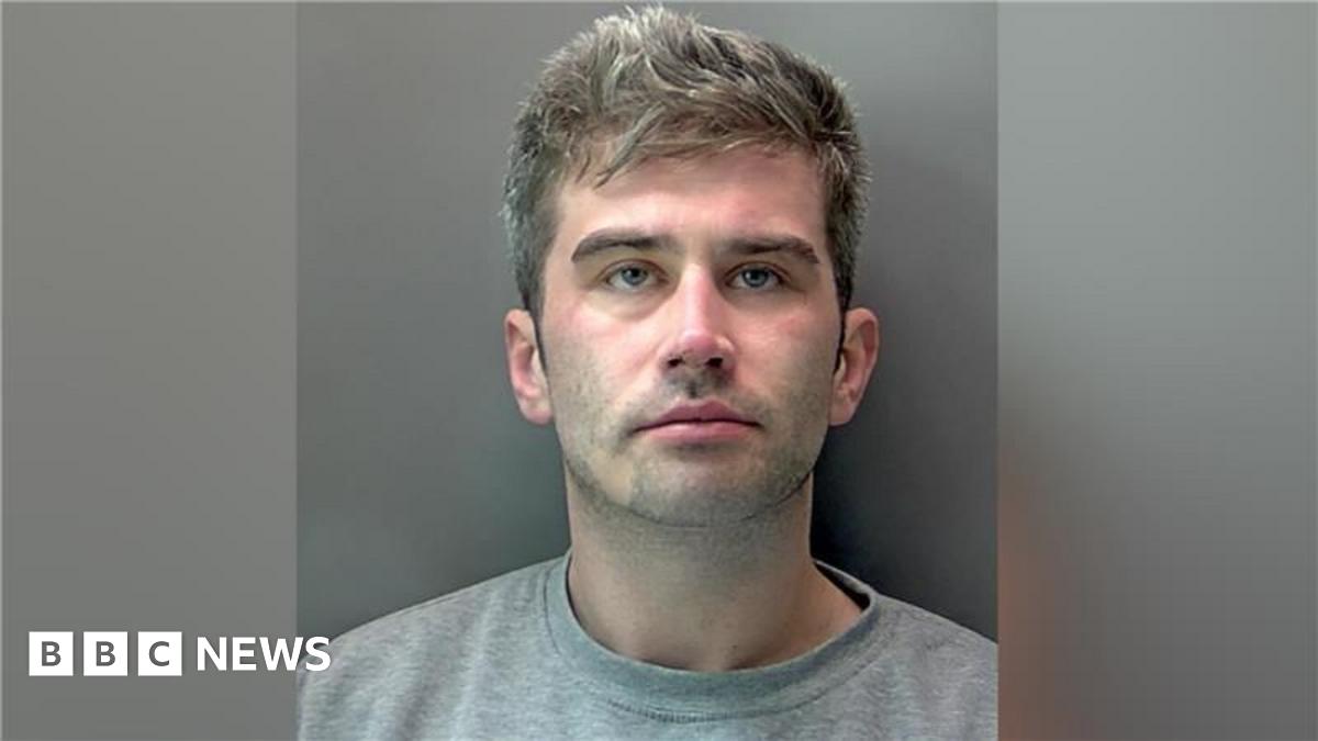 Robert Tyler-Jones standing on a bridge, in front of a canal and buildings, smiling at the camera, wearing a black cap, black top, trousers and a bag across him. He is standing next to a plant display.