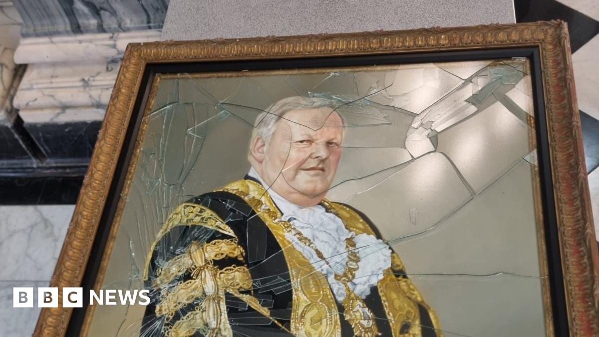 A photo of the damage to a gold-framed oil painting of former Belfast lord mayor Lord Browne.   The painting shows a grey-haired man in a black and gold ceremonial robe, wearing a gold mayoral chain around his neck.   Most of the glass protecting the painting has been shattered.