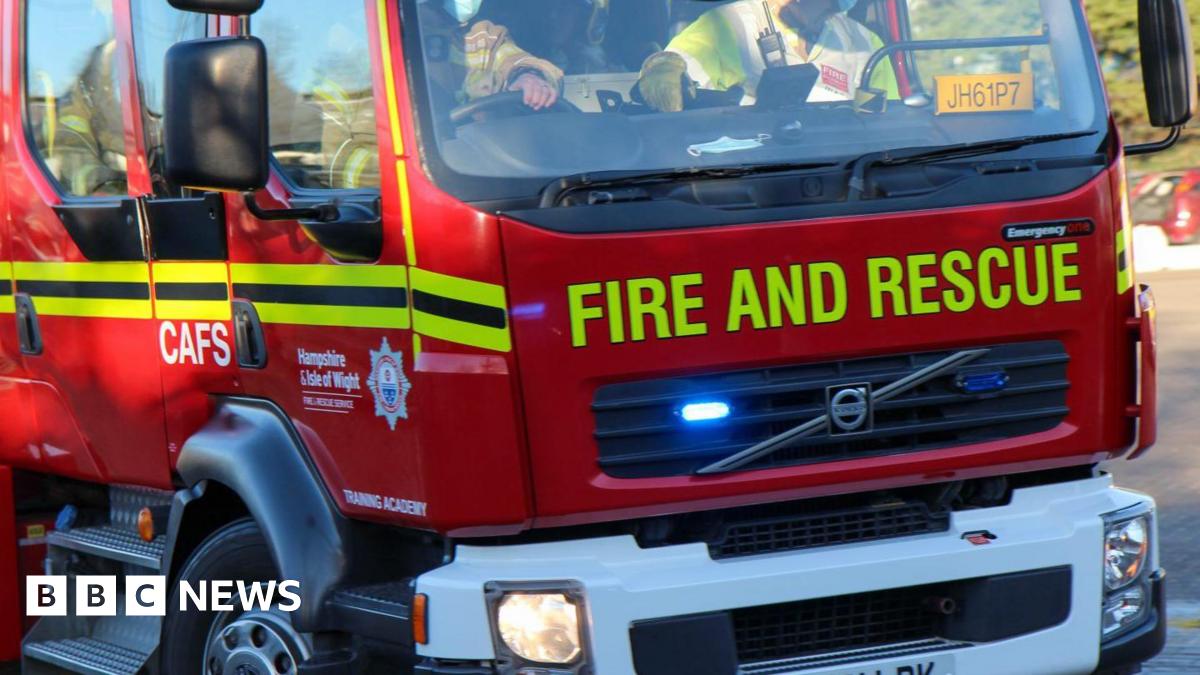 A Hampshire and Isle of Wight Fire and Rescue vehicle making a turn. "training academy is written on the driver's door. Two Fire and Rescue staff can bee seen in the front of the cab. The passenger is wearing a high-vis top over fire service overalls.