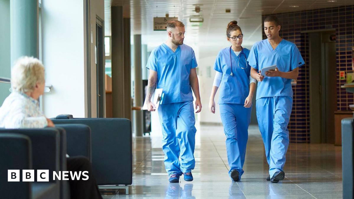 Three medics in blue scrubs, two men and a woman, walk along a hospital corridor. One of the men is holding a clipboard, the other some papers and the woman has a stethoscope around her neck. They are walking side by side, engaged in conversation. In the foreground, there is woman sitting on a black cushioned chair. She is turned away from the camera, looking towards the medics.