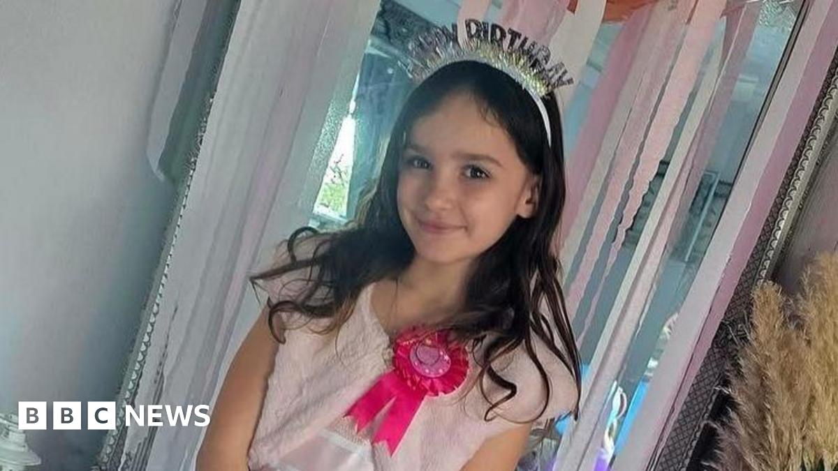 Aria smiles at the camera. She is wearing a pink birthday dress, a bright pink rosette and a birthday crown. She has long brown hair and the doorway behind her is decorated with pink streamers.