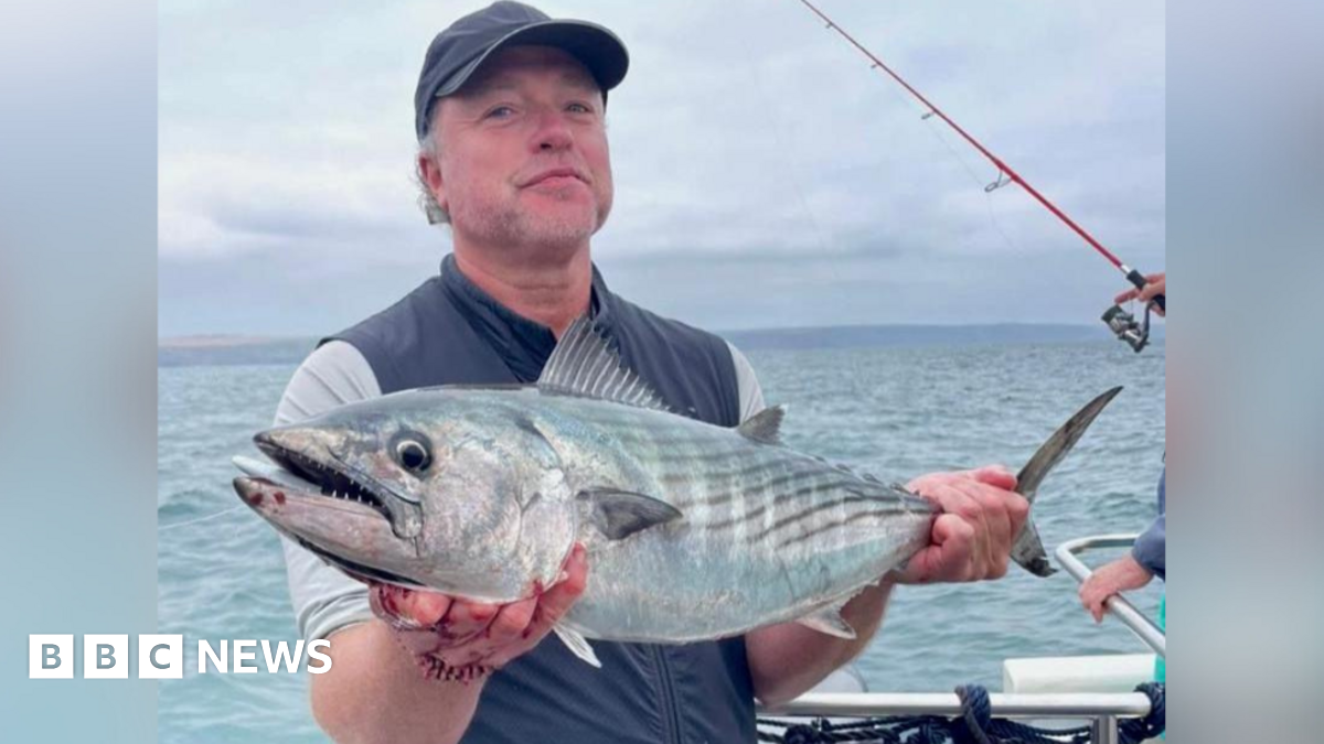A young woman stands on the left holding a fishing rod. She is smiling because she just caught a fish. A teenage boy stands on the right removing the fish from the hook. They are both wearing red life jackets.