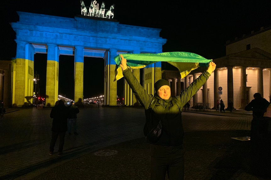 A man holds up a pro-Ukraine scarf, in front of the Brandenburg Gate lit up in the colours of Ukraine flag.