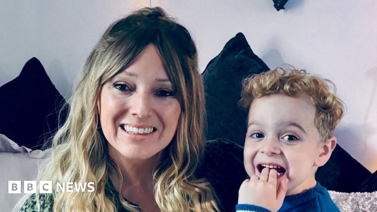 Vicky Ogg is pictured holding her son Kacey. They are both smiling. Vicky has long, wavy, blonde hair which has a fringe that is parted in the middle. Her son - who has curly, blonde hair - has two fingers in his mouth. They are sitting on a dark blue velvet sofa.