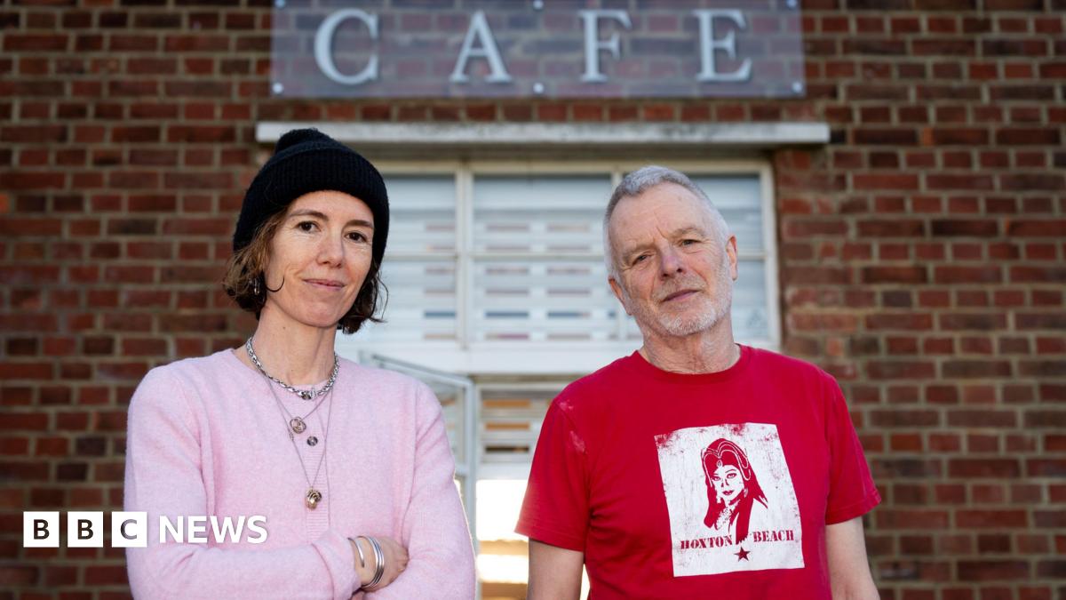 Emma Fernandez and Patrick Matthews standing in front of the lido cafe. Emma wears a pink cardigan and a black beanie hat, and Patrick wears a red t-shirt.